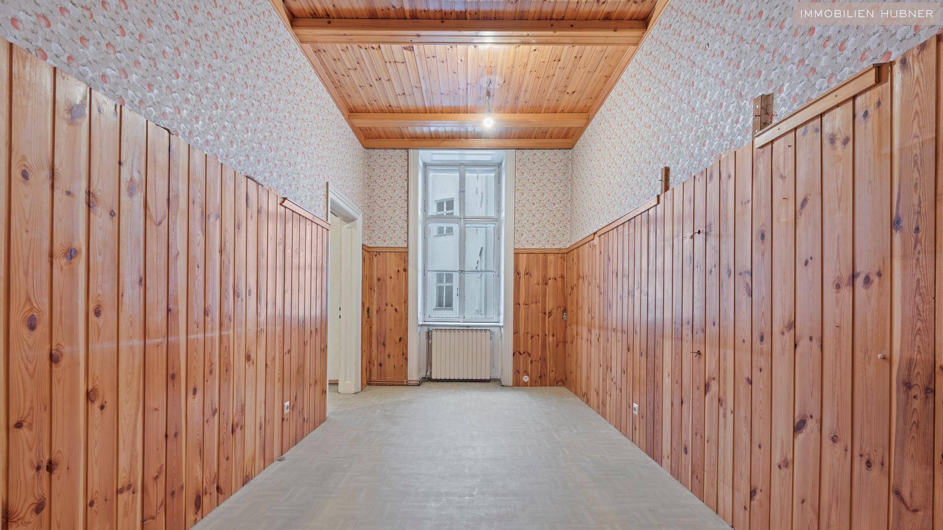 Langer Raum mit Holzvertäfelung, gemusterter Tapete und Fenster, der auf eine neue Nutzung wartet.