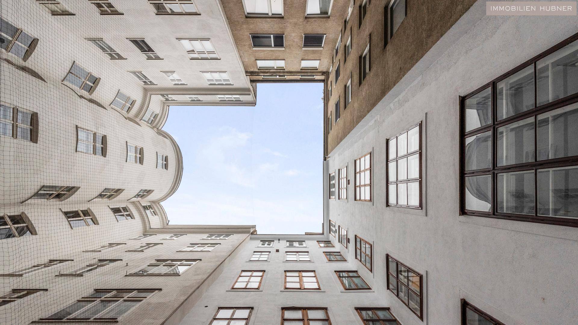 Blick nach oben in einen Innenhof, umgeben von hohen Gebäudefassaden mit vielen Fenstern.