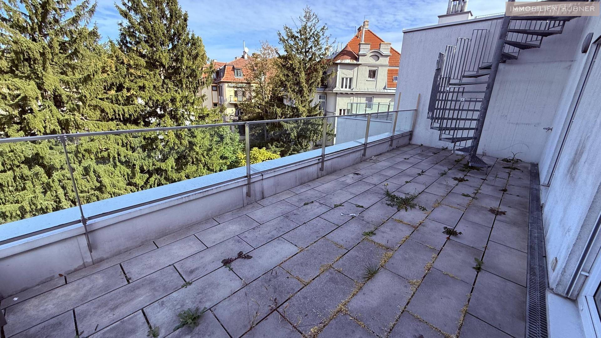 Terrasse mit Steinplatten, Glasgeländer und einer Wendeltreppe, umgeben von hohen Bäumen.