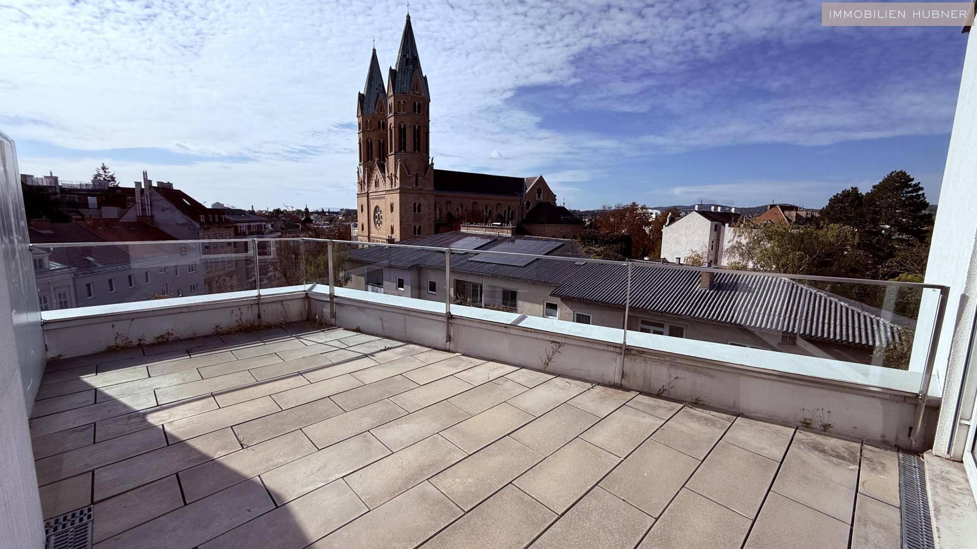 Geräumige Terrasse mit Steinplatten, Glasgeländer und weitem Blick auf die Stadt und eine Kirche.