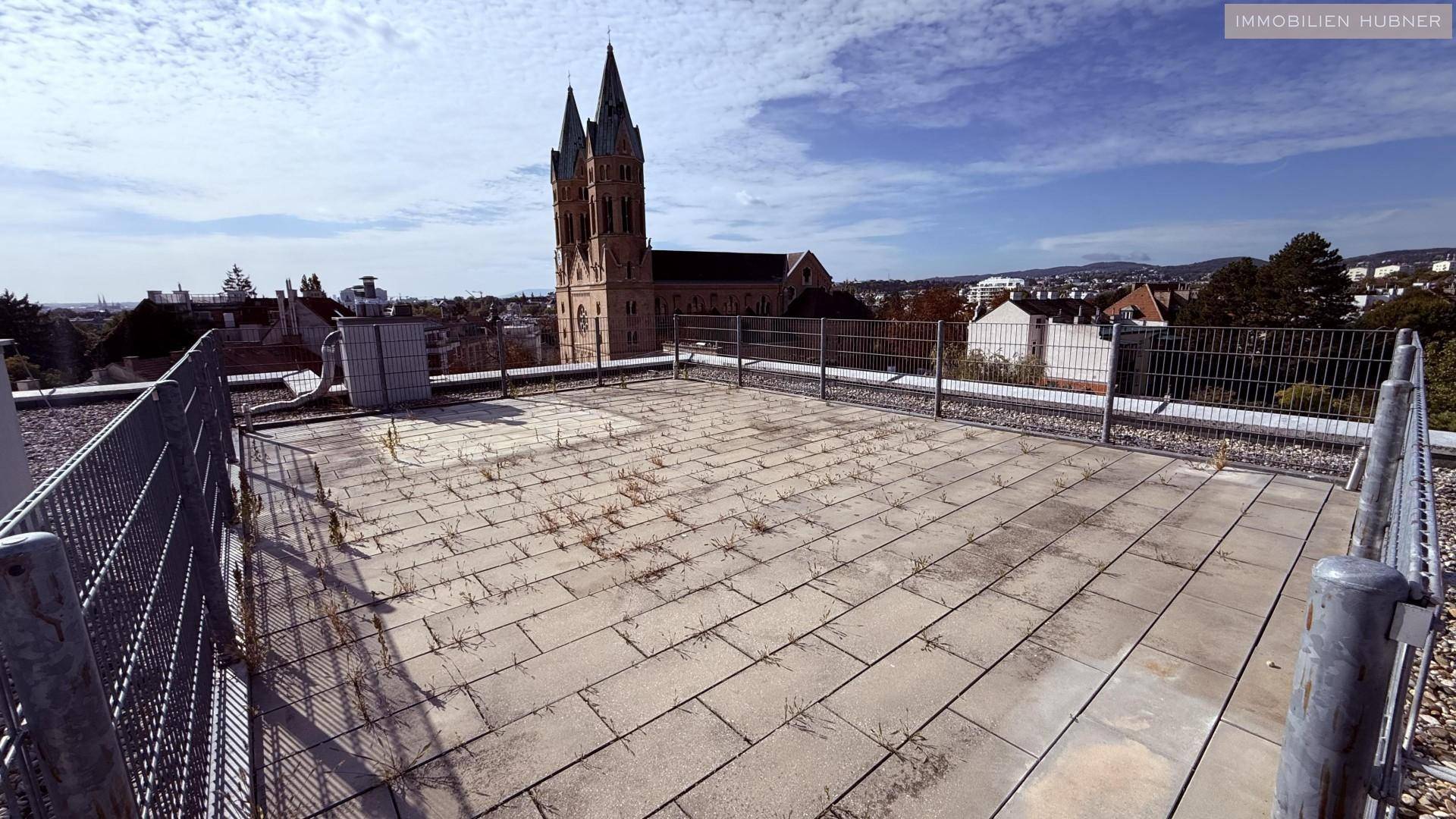 Große Dachterrasse mit Steinplatten und Metallzaun, bietet einen weiten Blick auf die Umgebung und eine Kirche.