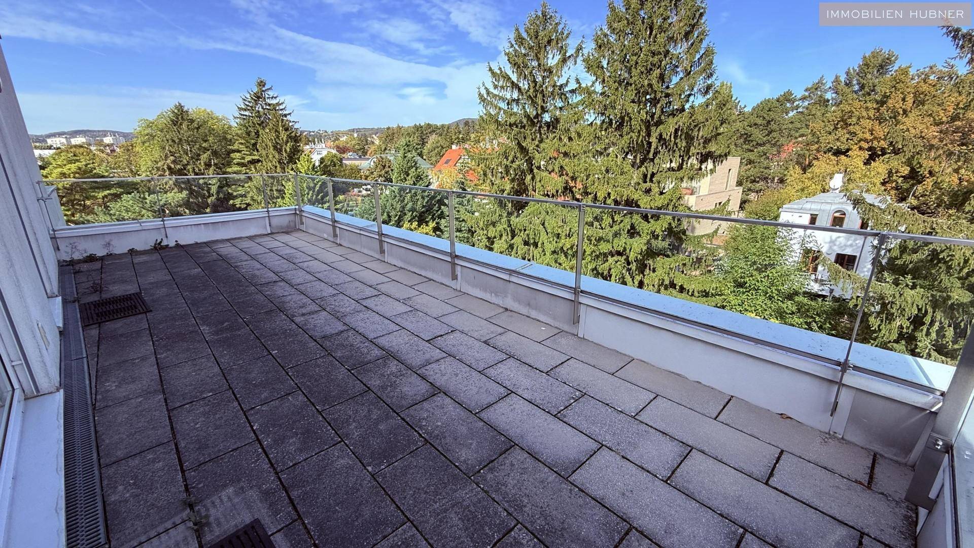 Geräumige Terrasse mit Fliesenboden und Glasgeländer, umgeben von viel Grün und Bäumen.