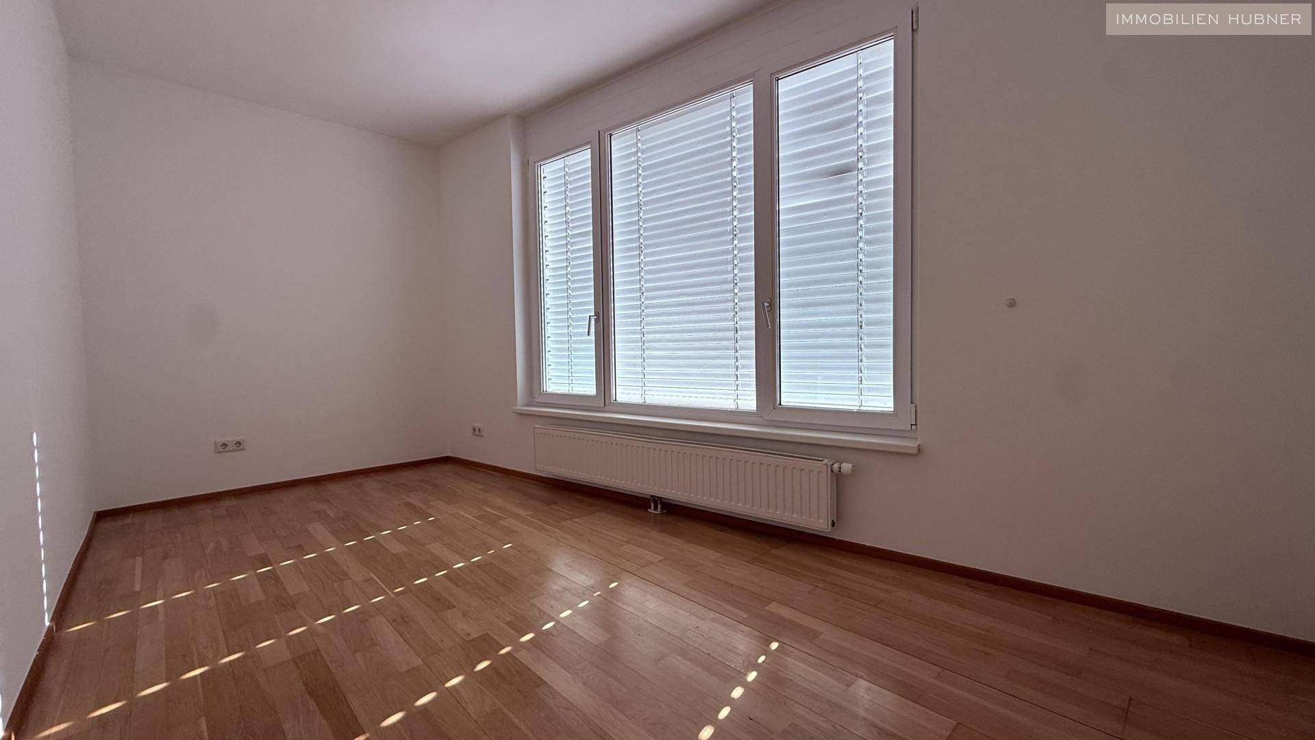 Helles Zimmer mit Holzboden und breitem Fenster, ausgestattet mit Jalousien für Privatsphäre.