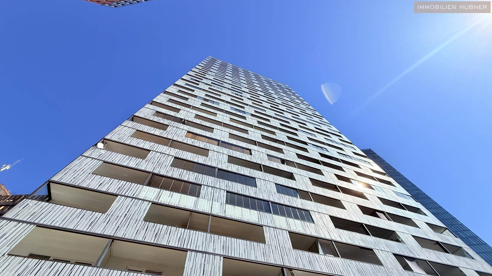 Hohes modernes Gebäude mit strukturierter weißer Fassade und zahlreichen Fenstern unter blauem Himmel.