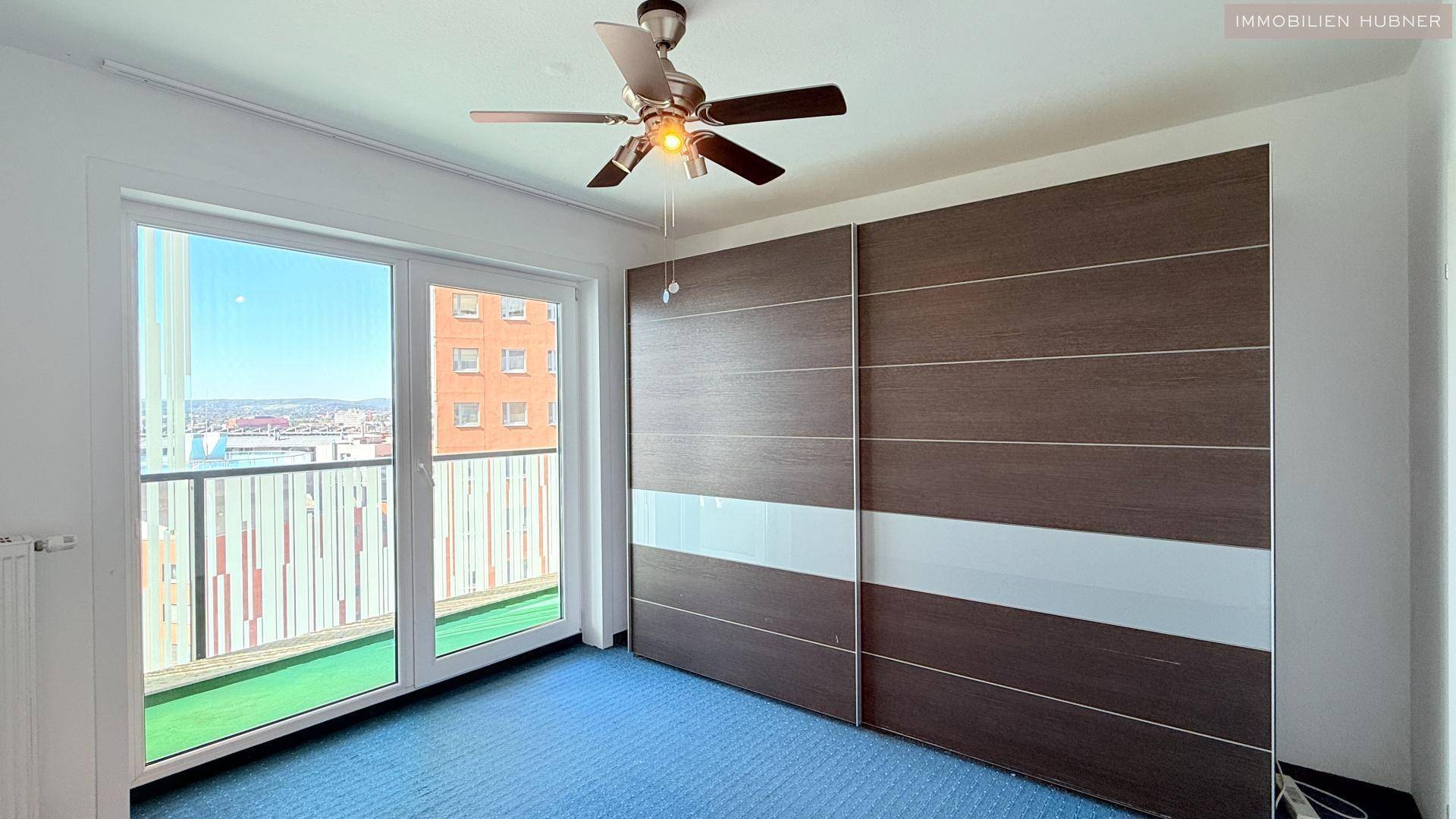 Schlafzimmer mit blauem Teppich, großem Schiebetürenschrank und Zugang zum Balkon.