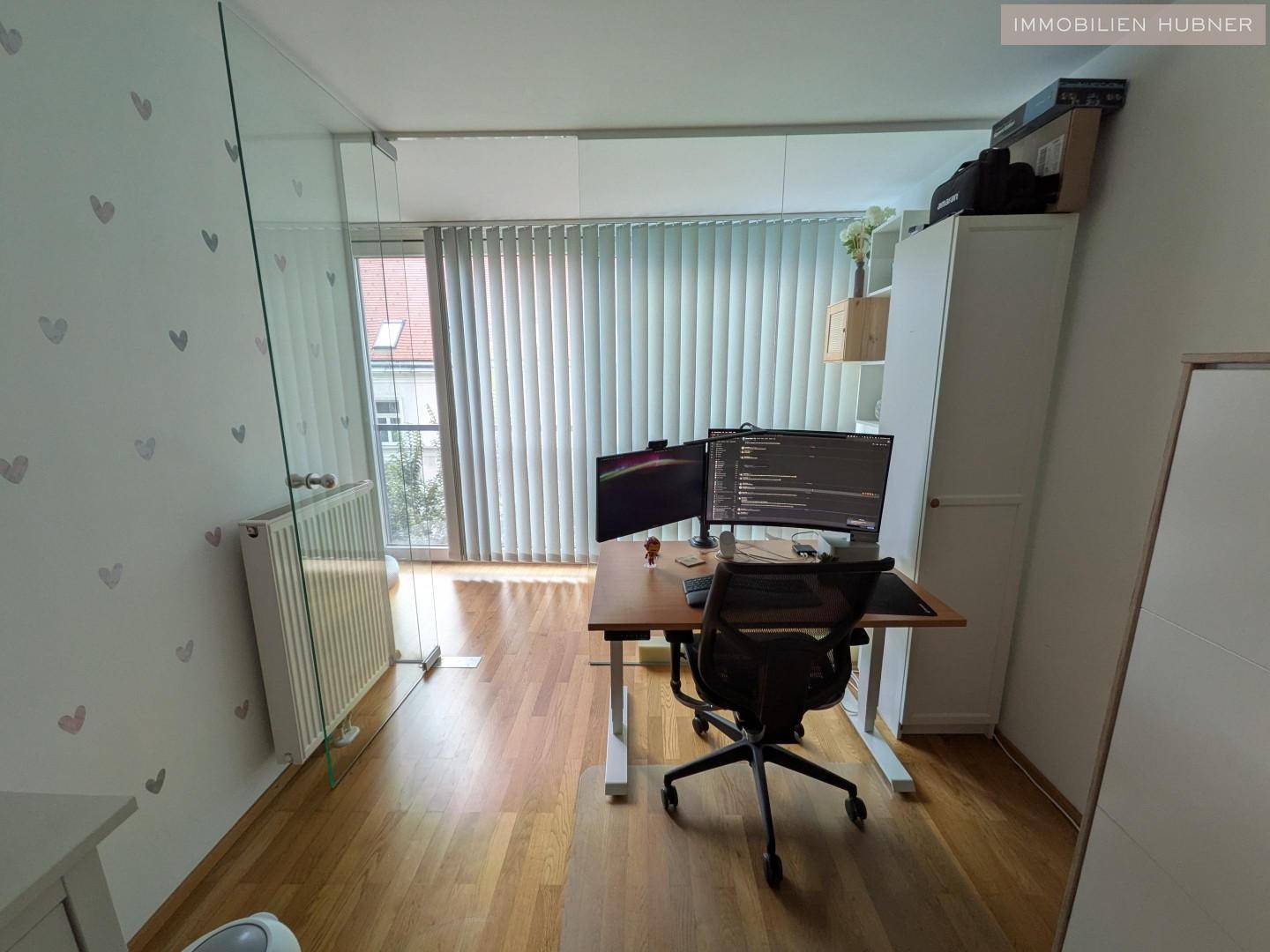 Helles Arbeitszimmer mit großem Schreibtisch und Computermonitoren, abgetrennt durch eine Glaswand.
