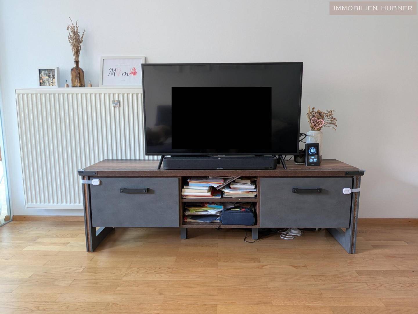 Detailansicht des Wohnbereichs mit modernem Fernseher auf einem Holz-Metall-Lowboard und Heizkörper.