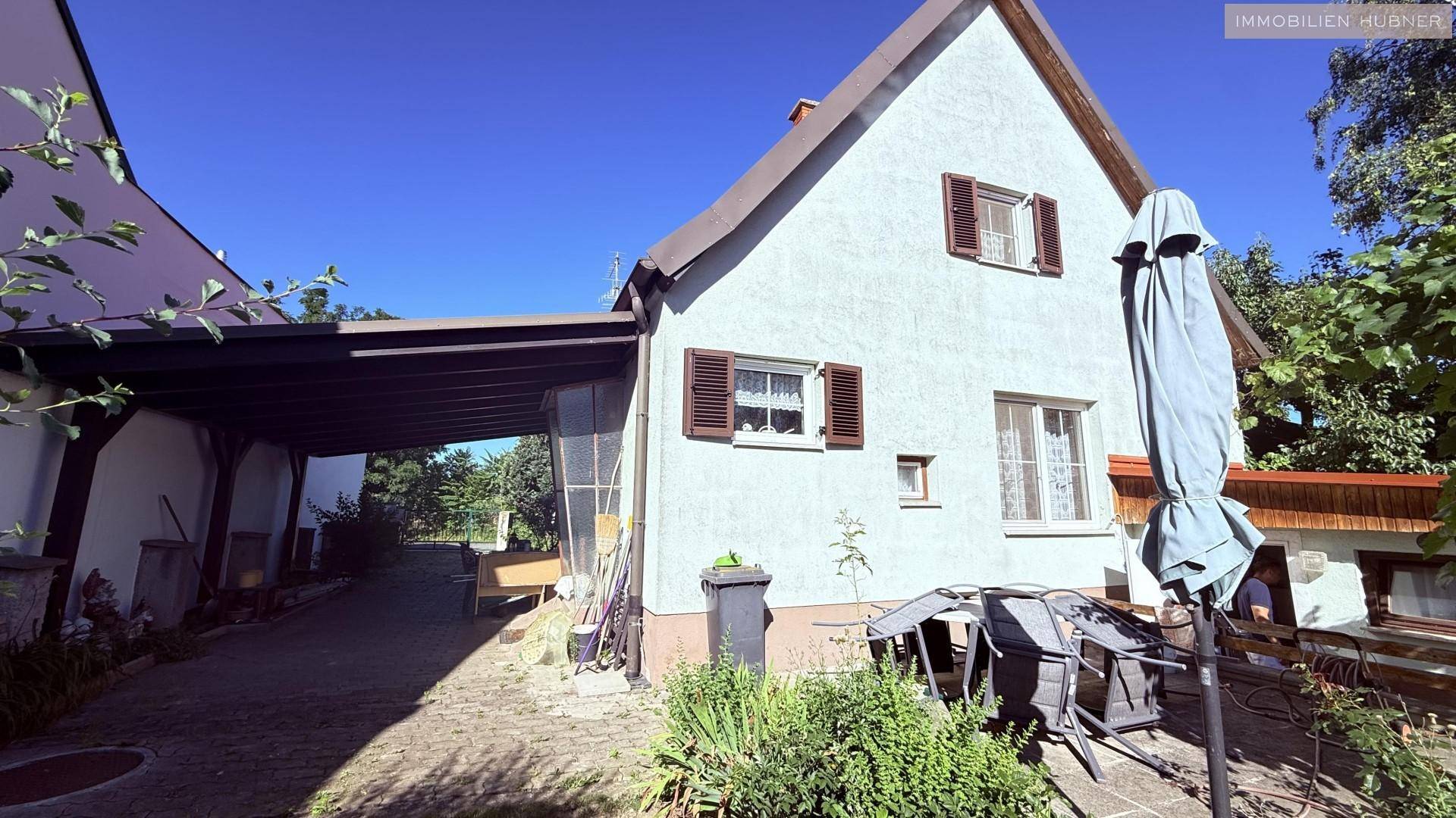 Hausseite mit Carport, gepflastertem Bereich und einer möblierten Sitzecke im Freien.