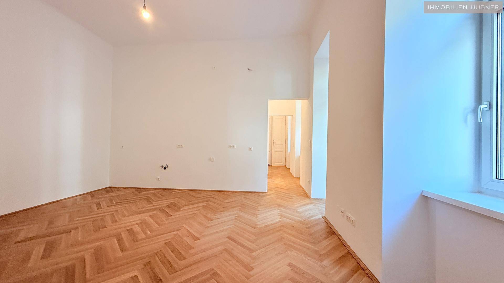 Heller Wohnbereich mit Fischgrätparkett und einem Fenster, das viel Tageslicht hereinlässt.