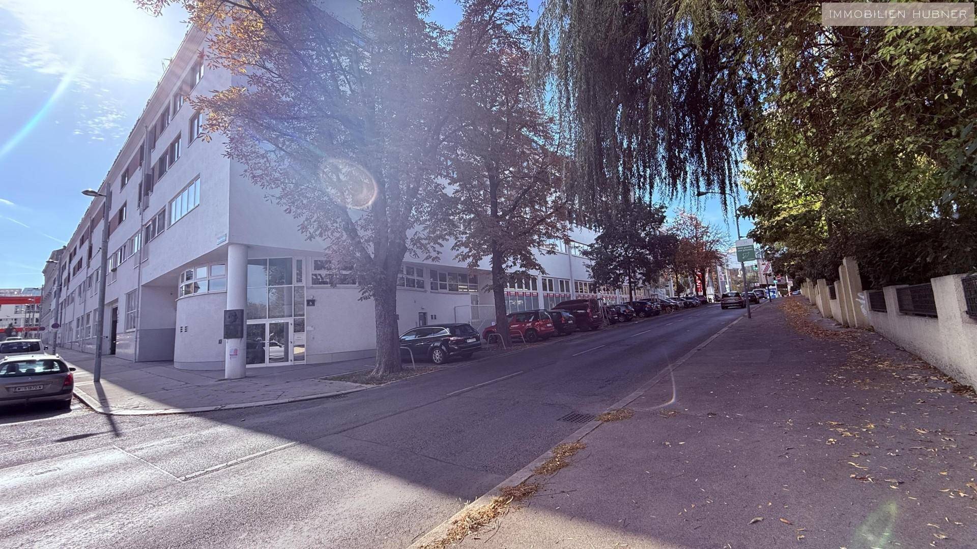 Gebäudeansicht von der Straße mit Bäumen und parkenden Autos, sonniger Tag.