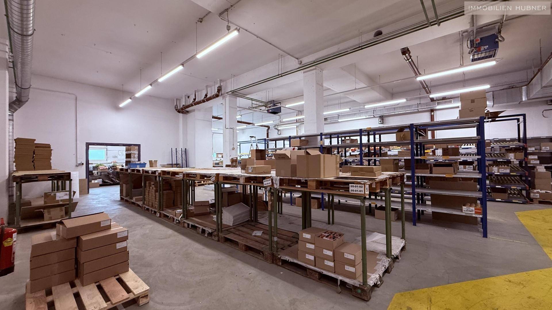 Großer Lagerraum mit Arbeitstischen, Paletten und Kartons, gut beleuchtet.