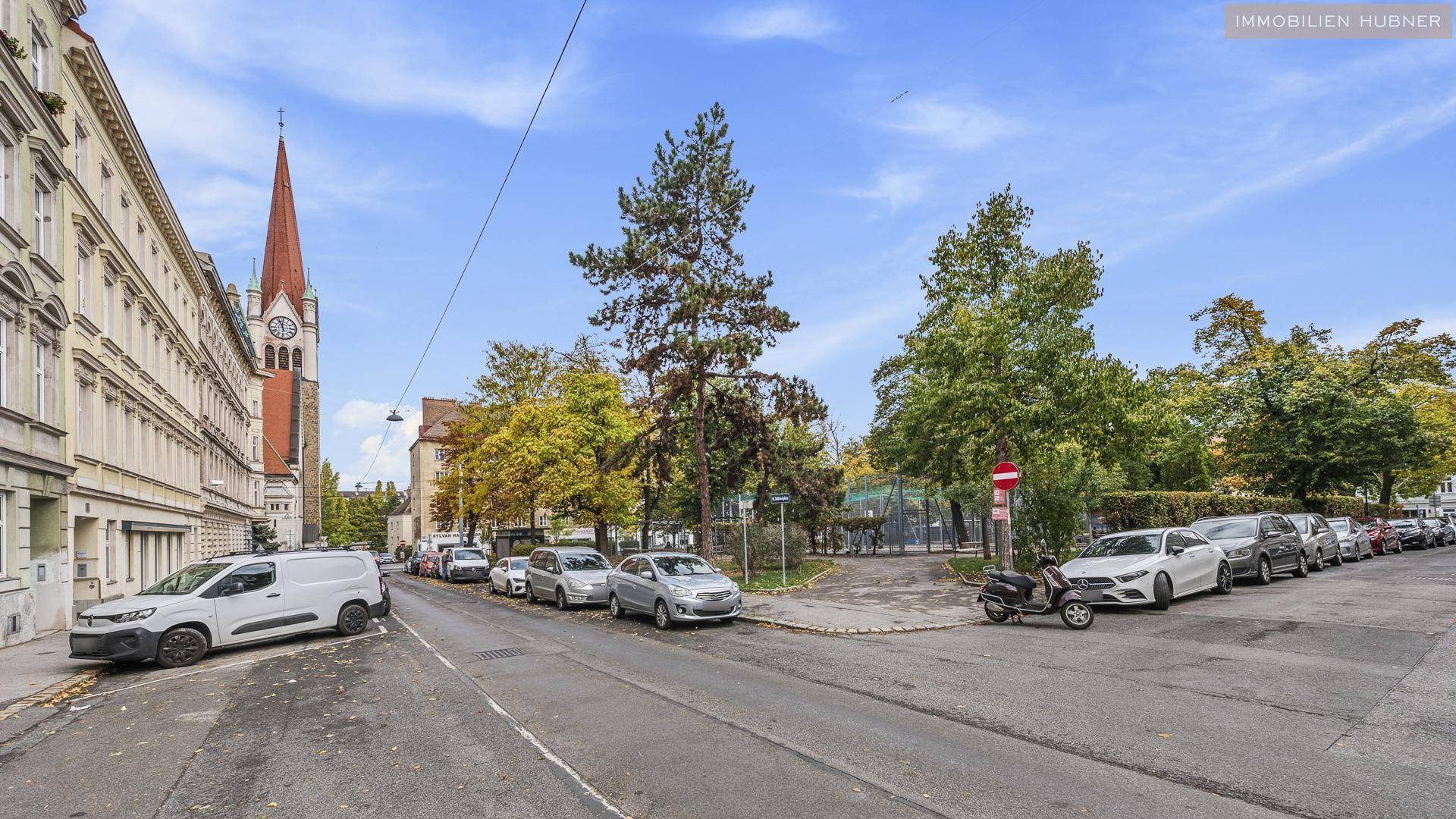Stadtstraße mit parkenden Autos, Bäumen und einer Kirche im Hintergrund unter blauem Himmel.