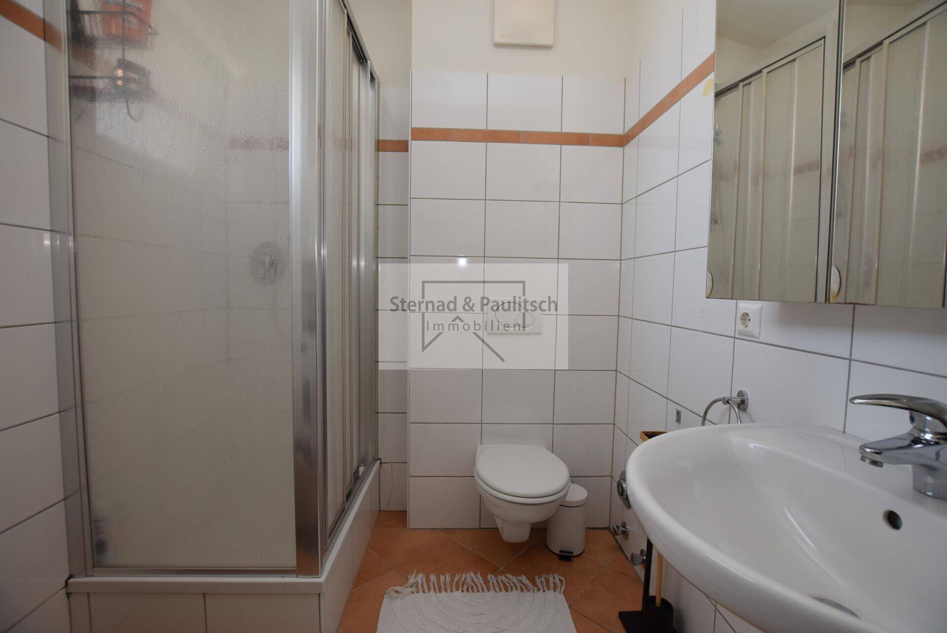 Badezimmer mit Duschkabine, wandhängendem WC und Waschbecken, komplett gefliest.