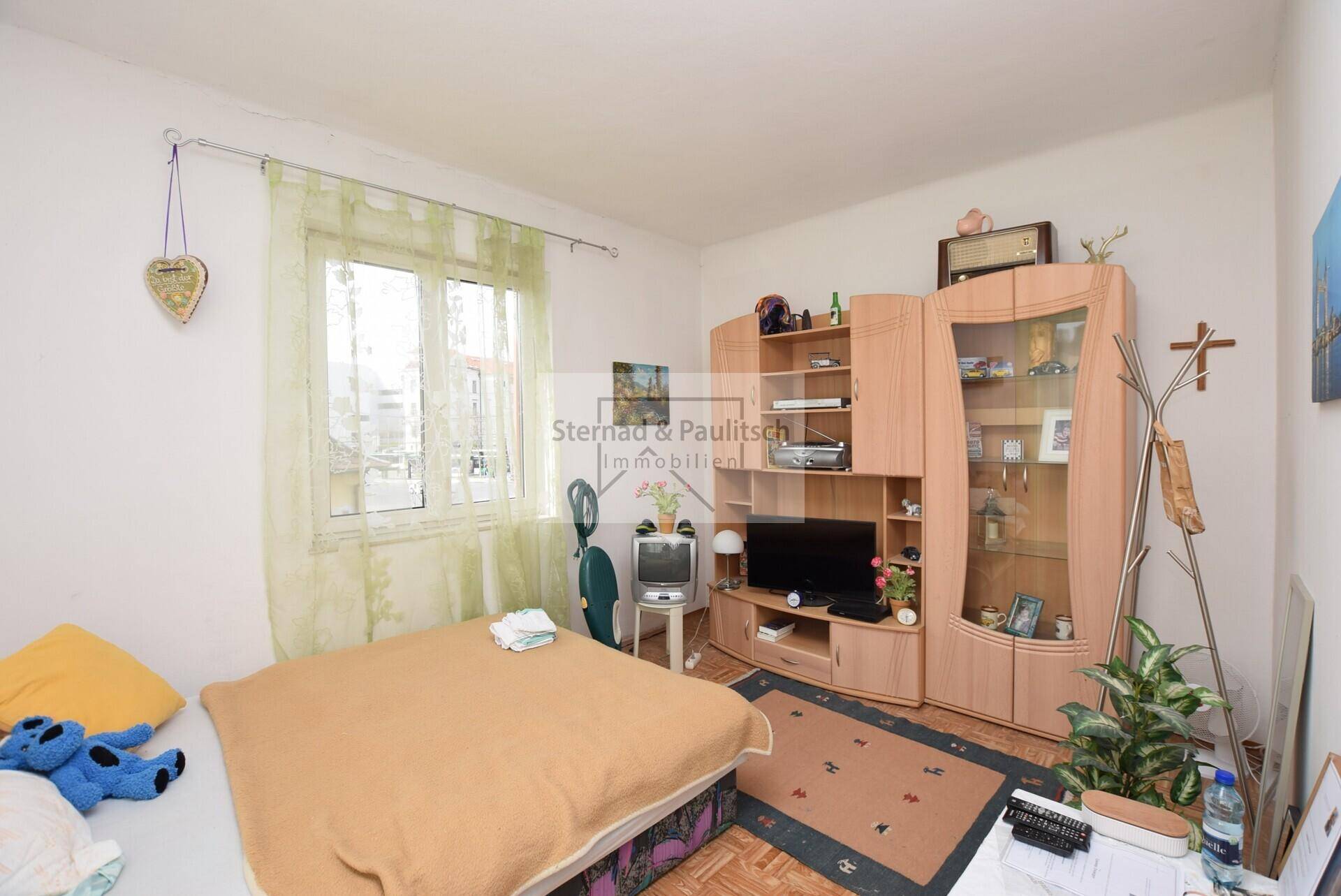 Schlafzimmer mit Bett, Schrankwand und Fenster mit hellem Vorhang.