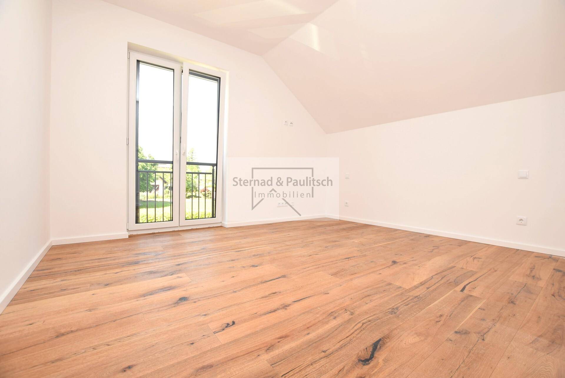 Geräumiges Zimmer mit Holzboden, Dachschräge und großen Fenstern mit Blick ins Grüne.