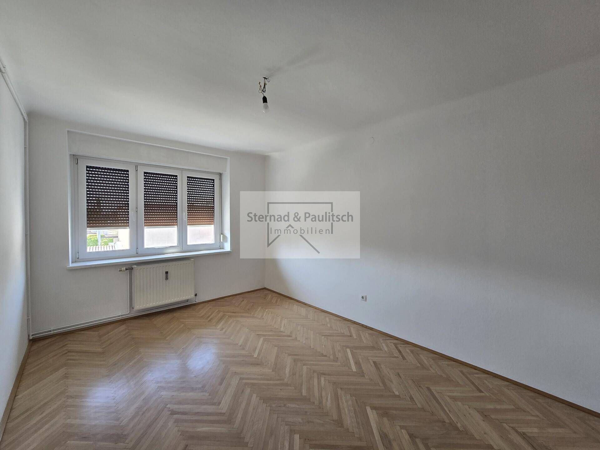 Lichtdurchflutetes Zimmer mit klassischem Fischgrätparkett und großem Fenster mit Jalousien.