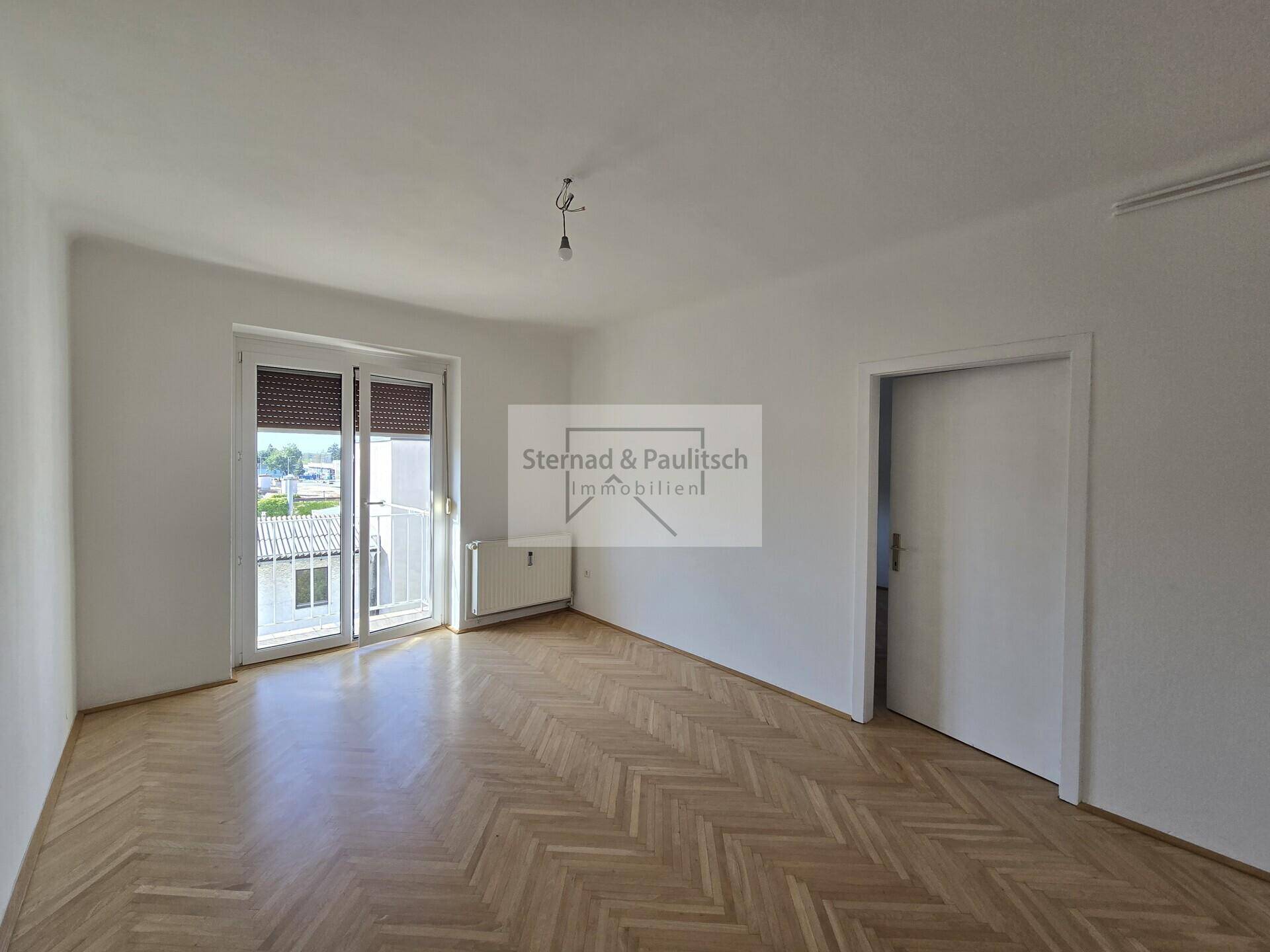 Helles Zimmer mit Fischgrätparkett und einem französischen Balkon, der einen Blick nach draußen bietet.