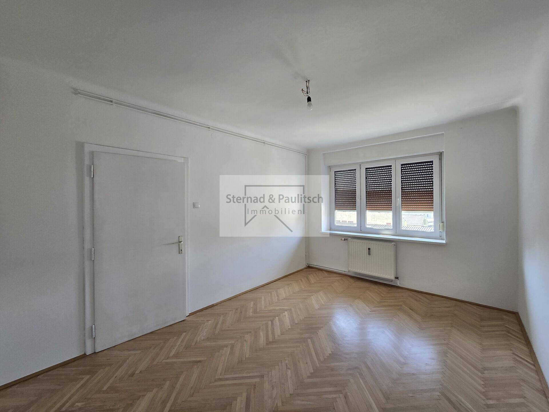 Heller Raum mit Fischgrätparkettboden, Fenster mit Jalousien und einer weißen Tür.