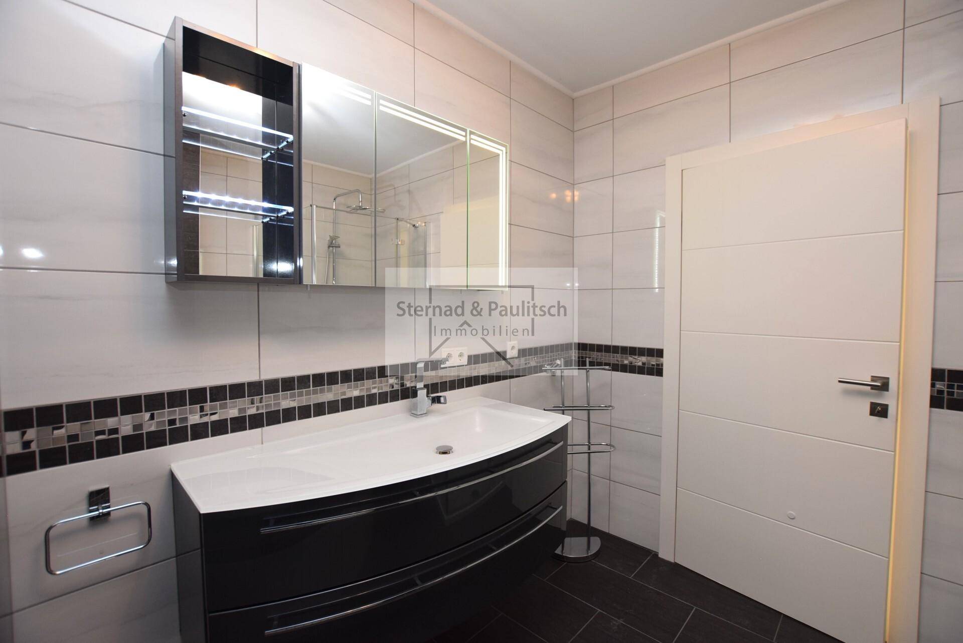 Elegantes Badezimmer mit modernem Waschtisch, Spiegelschrank und dunklen Bodenfliesen.