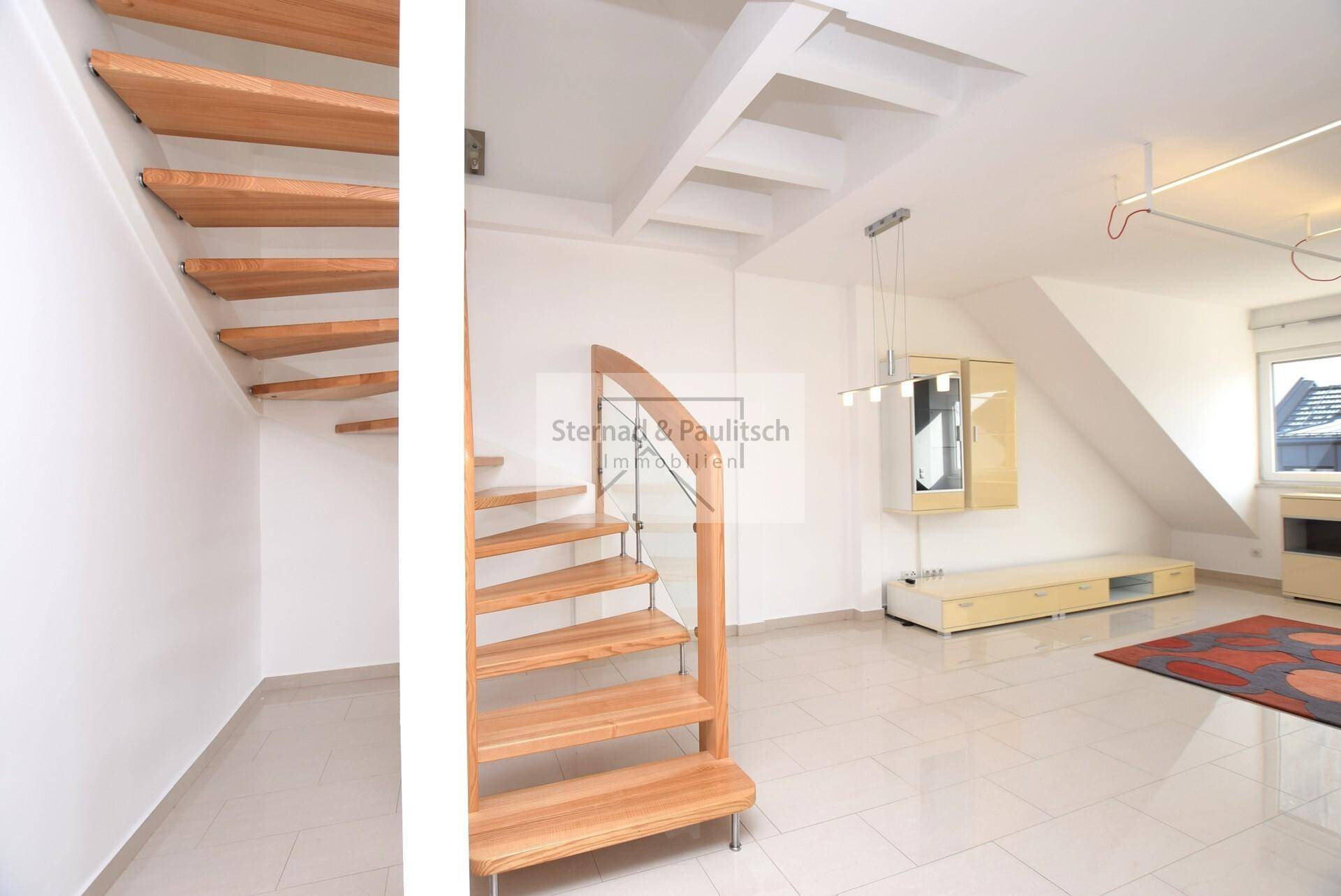 Offener Wohnbereich mit moderner Holztreppe und Fliesenboden unter schrägen Dachflächen.
