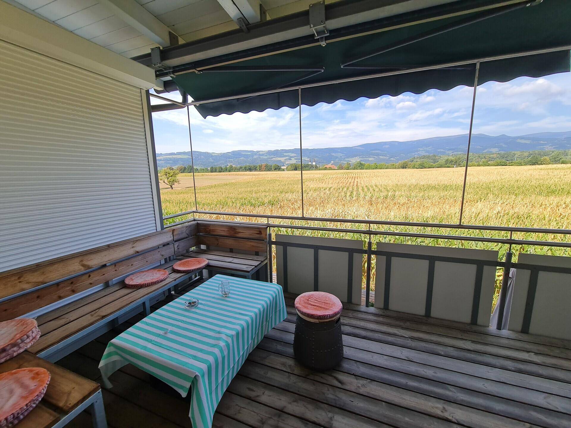 Überdachter Balkon mit Holzbänken und Tisch, bietet einen weiten, grünen Blick auf die Landschaft.