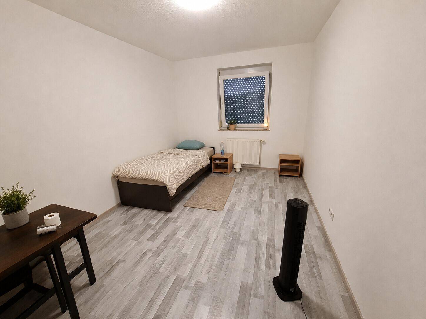 Helles Schlafzimmer mit Einzelbett, Laminatboden und einem modernen Standventilator für Komfort.