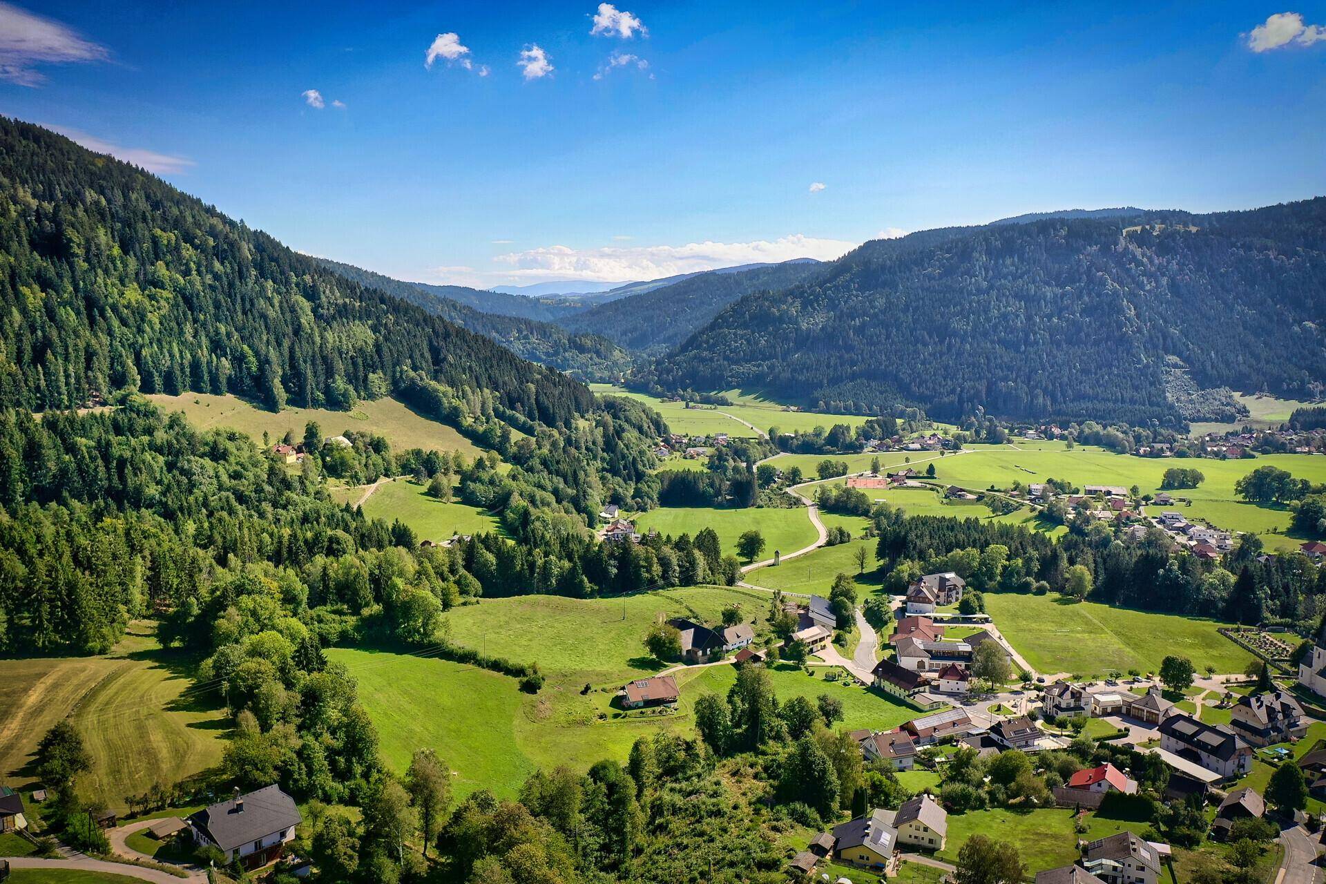 Weitläufige Luftaufnahme eines Tals mit einem Dorf, umgeben von grünen Hügeln und Wäldern unter blauem Himmel.