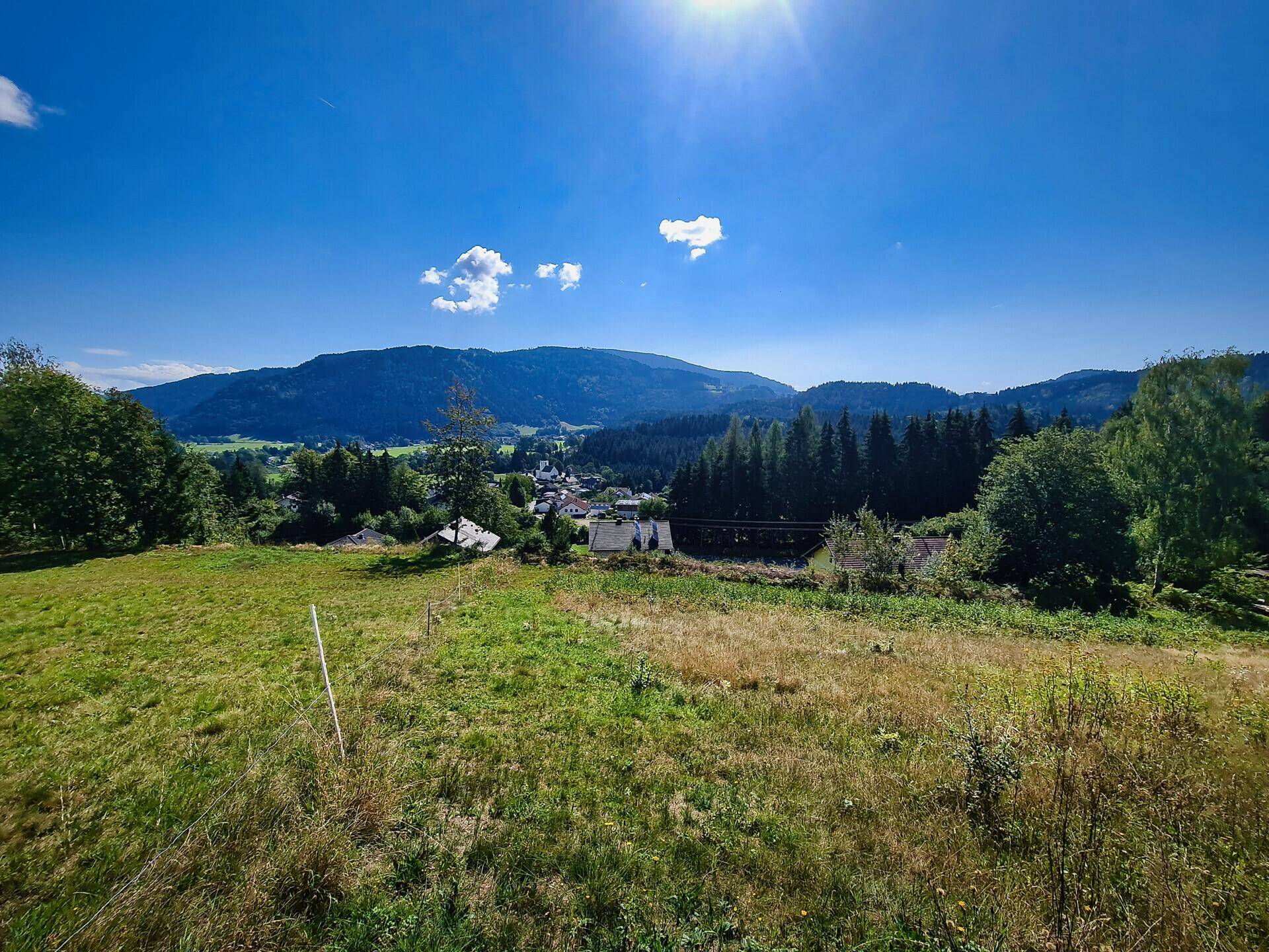 Weitläufiges grünes Feld im Vordergrund mit einem Dorf und bewaldeten Bergen im Hintergrund unter strahlend blauem Himmel.