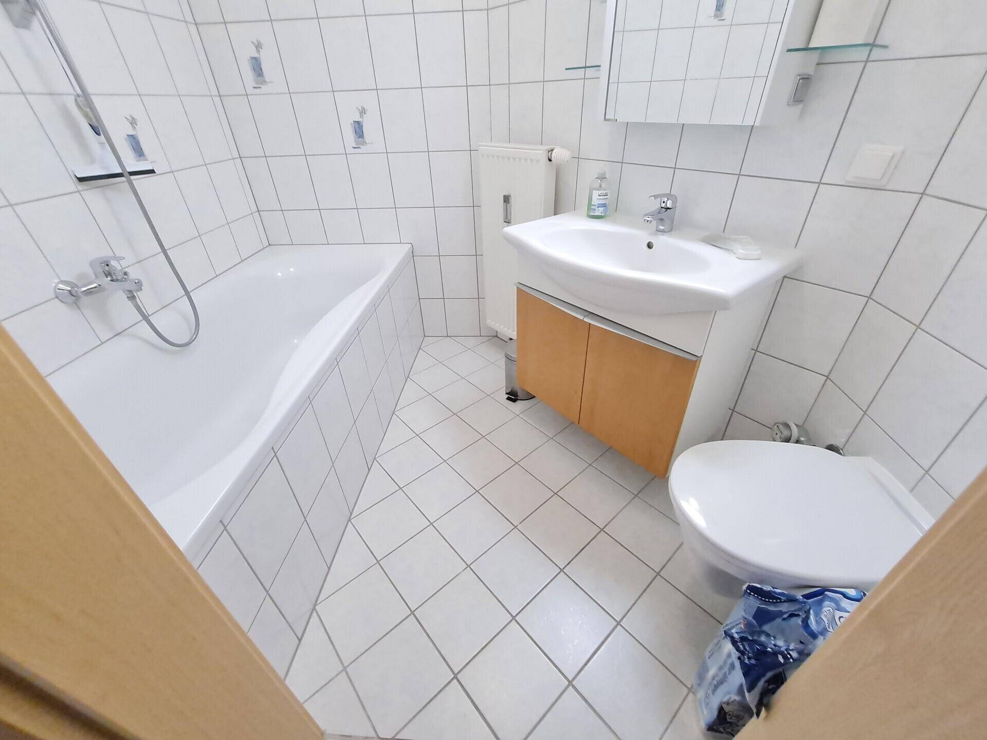 Helles Badezimmer mit weißen Fliesen, Badewanne, WC und einem Waschbecken mit Unterschrank.