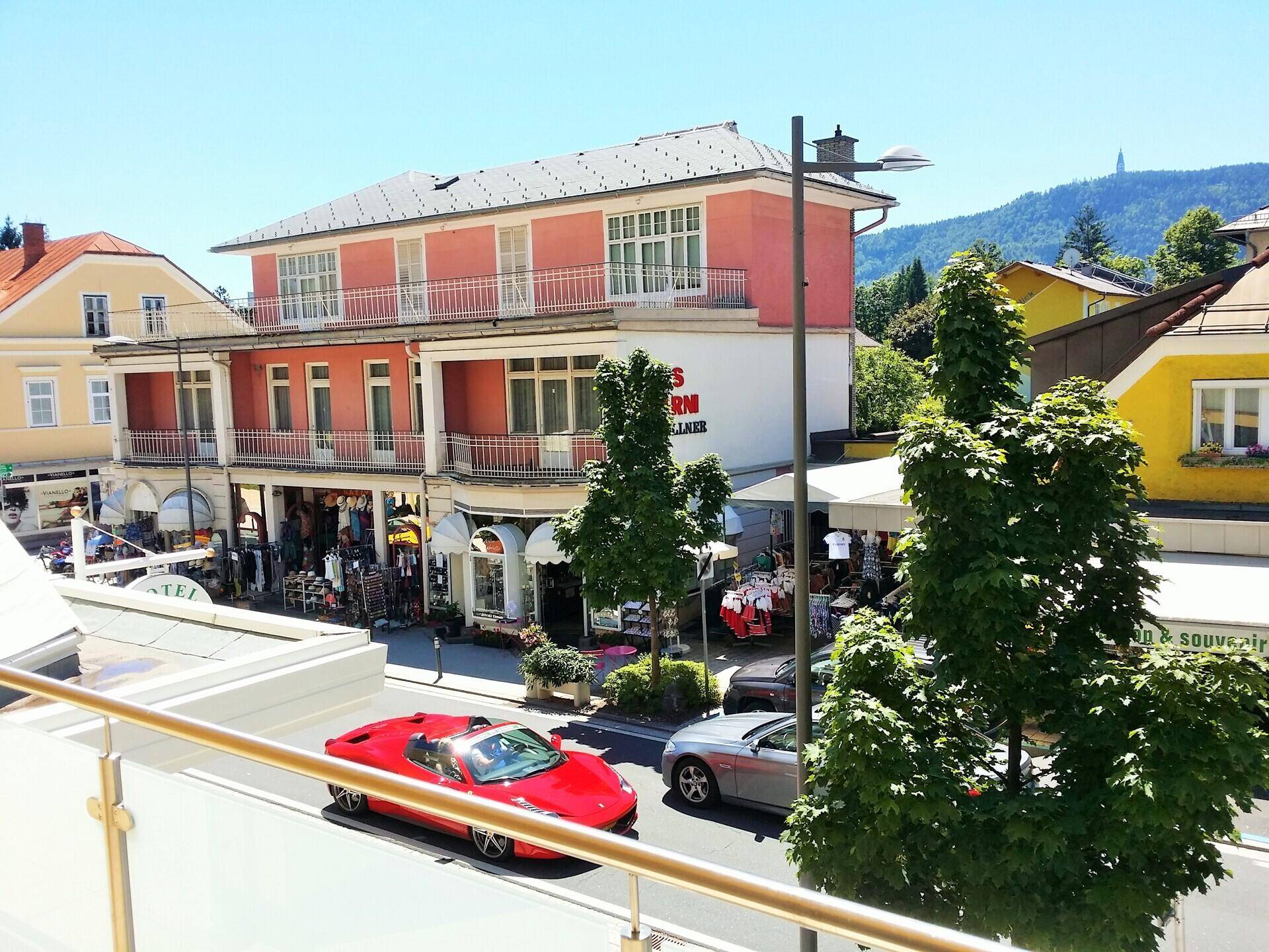 Balkonblick auf eine belebte Straße mit Geschäften und malerischer Bergkulisse im Hintergrund.