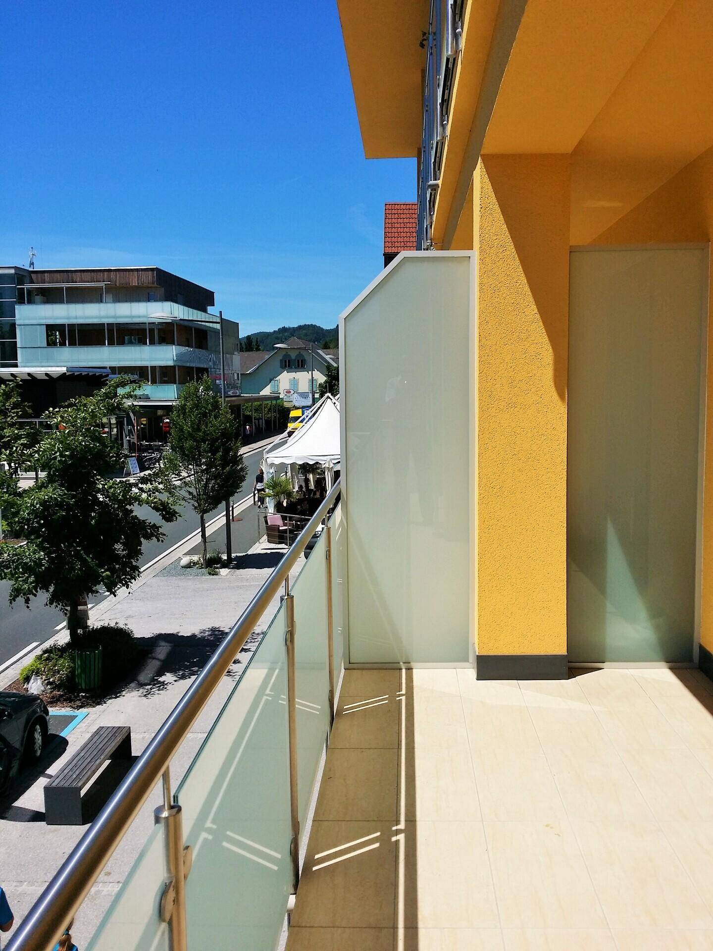 Heller Balkon mit Glasgeländer und Blick auf die belebte Straße und umliegende Gebäude.