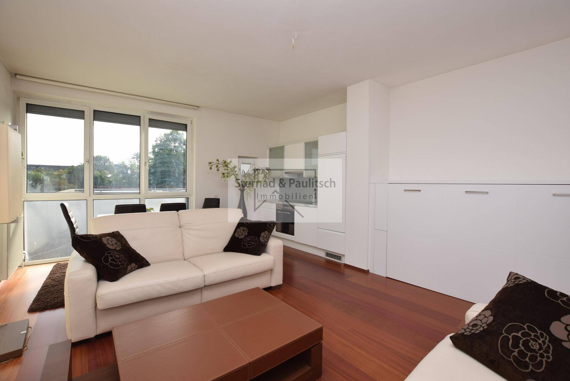 Wohnzimmer mit Sofa, Couchtisch und integriertem Schrankbett, hell durch große Fenster.