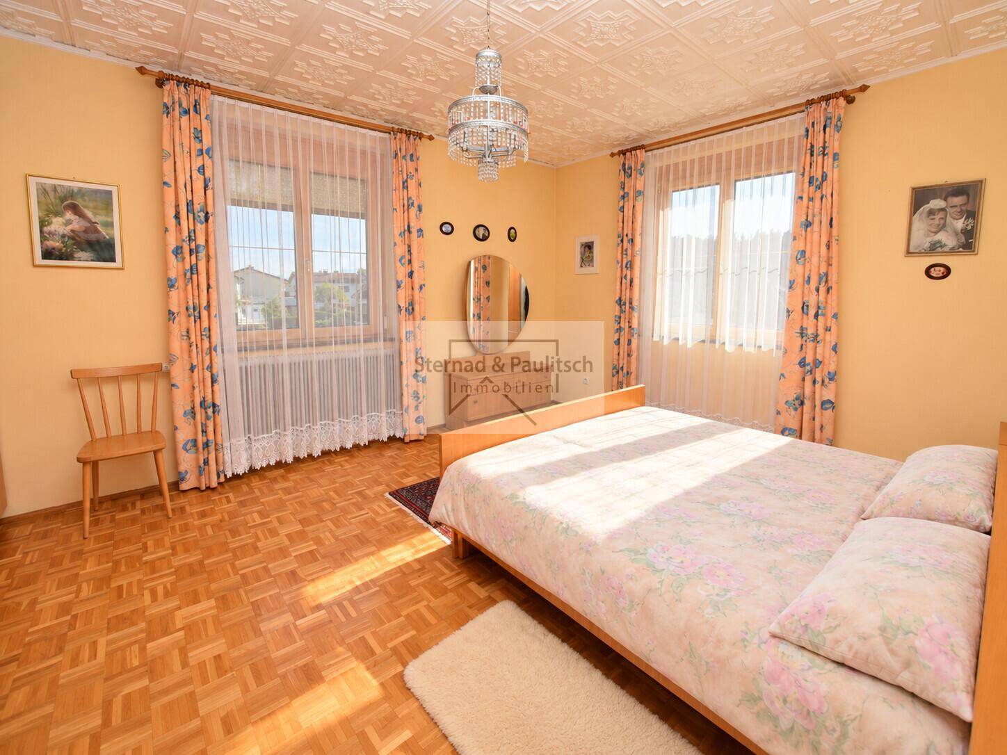 Helles Schlafzimmer mit Doppelbett, Parkettboden und zwei Fenstern für viel Tageslicht.