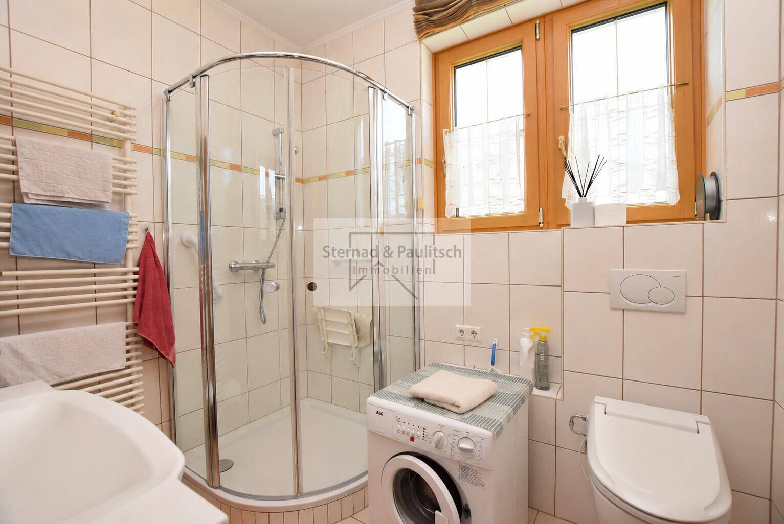 Badezimmer mit Duschkabine, WC, Waschbecken und Waschmaschine, hell gefliest.