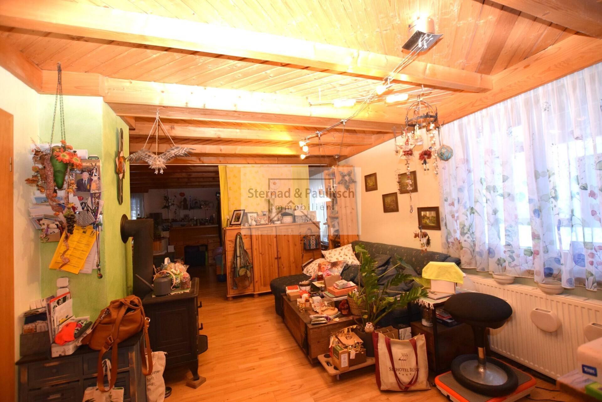 Wohnzimmer mit Holzbalkendecke, Holzofen und Fenster mit Vorhängen, gemütliche Atmosphäre.