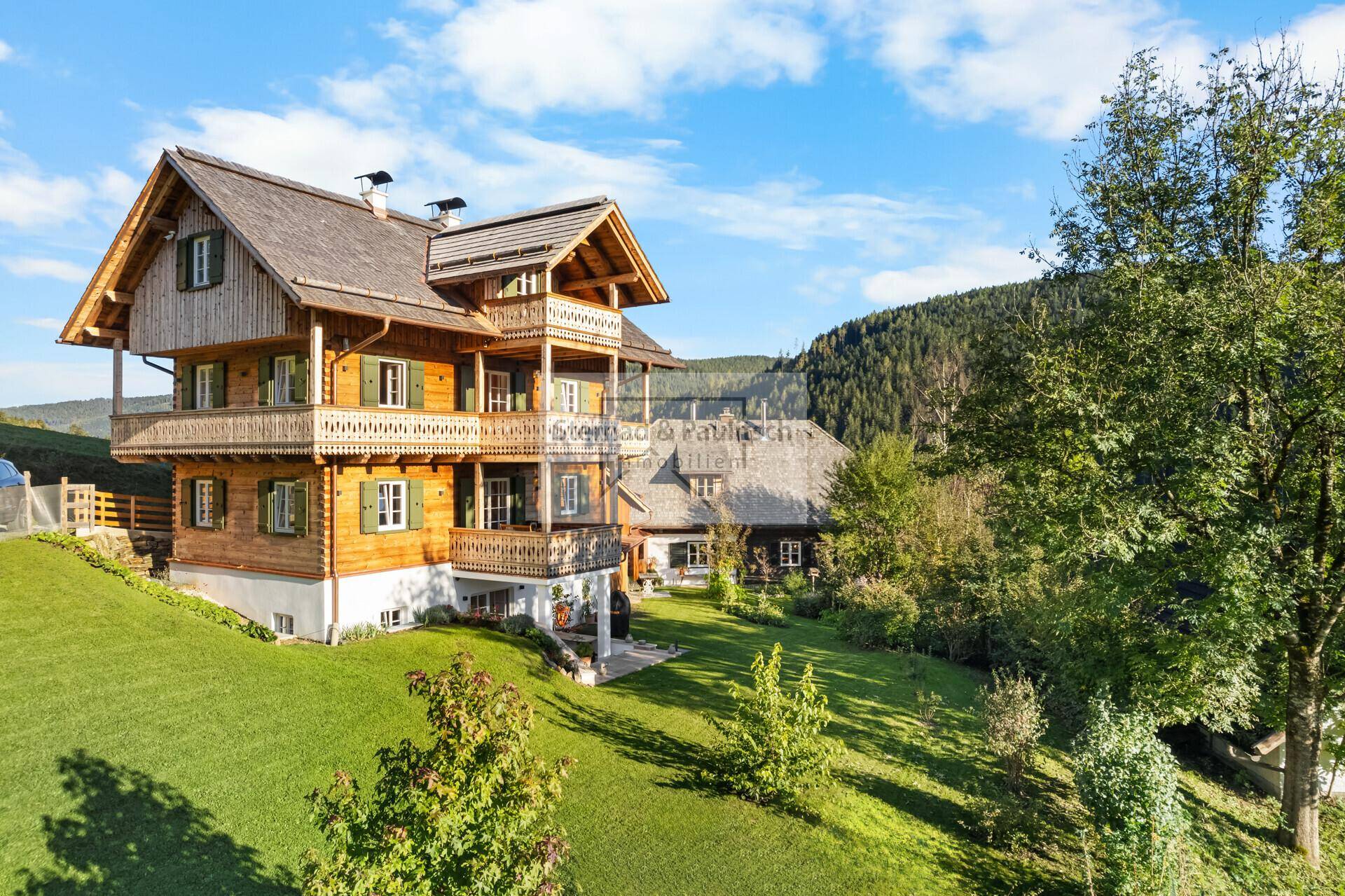 Traditionelles Holzchalet mit mehreren Balkonen und grüner Hanglage vor bewaldeten Bergen.