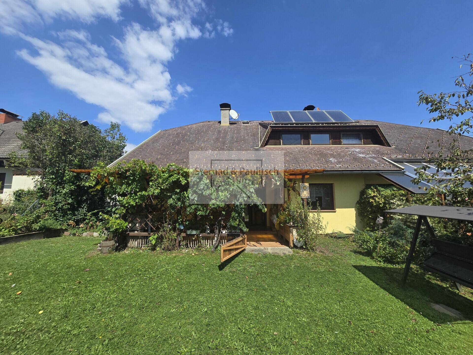 Sommerliche Ansicht des Hauses mit grünem Rasen, üppiger Bepflanzung und Solaranlage auf dem Dach.
