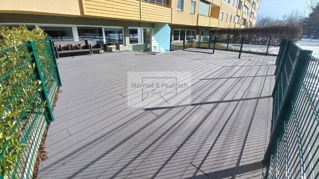 Große, gepflasterte Terrasse mit Blick auf das Gebäude, umgeben von einem Zaun und Hecken.