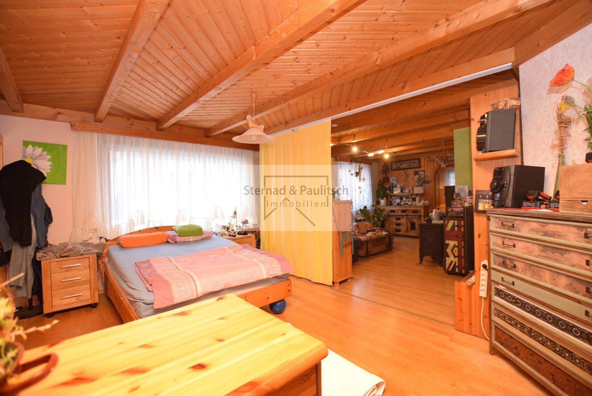 Offener Wohnbereich mit Holzbalkendecke und Holzboden, verbunden mit einem Schlafzimmer.