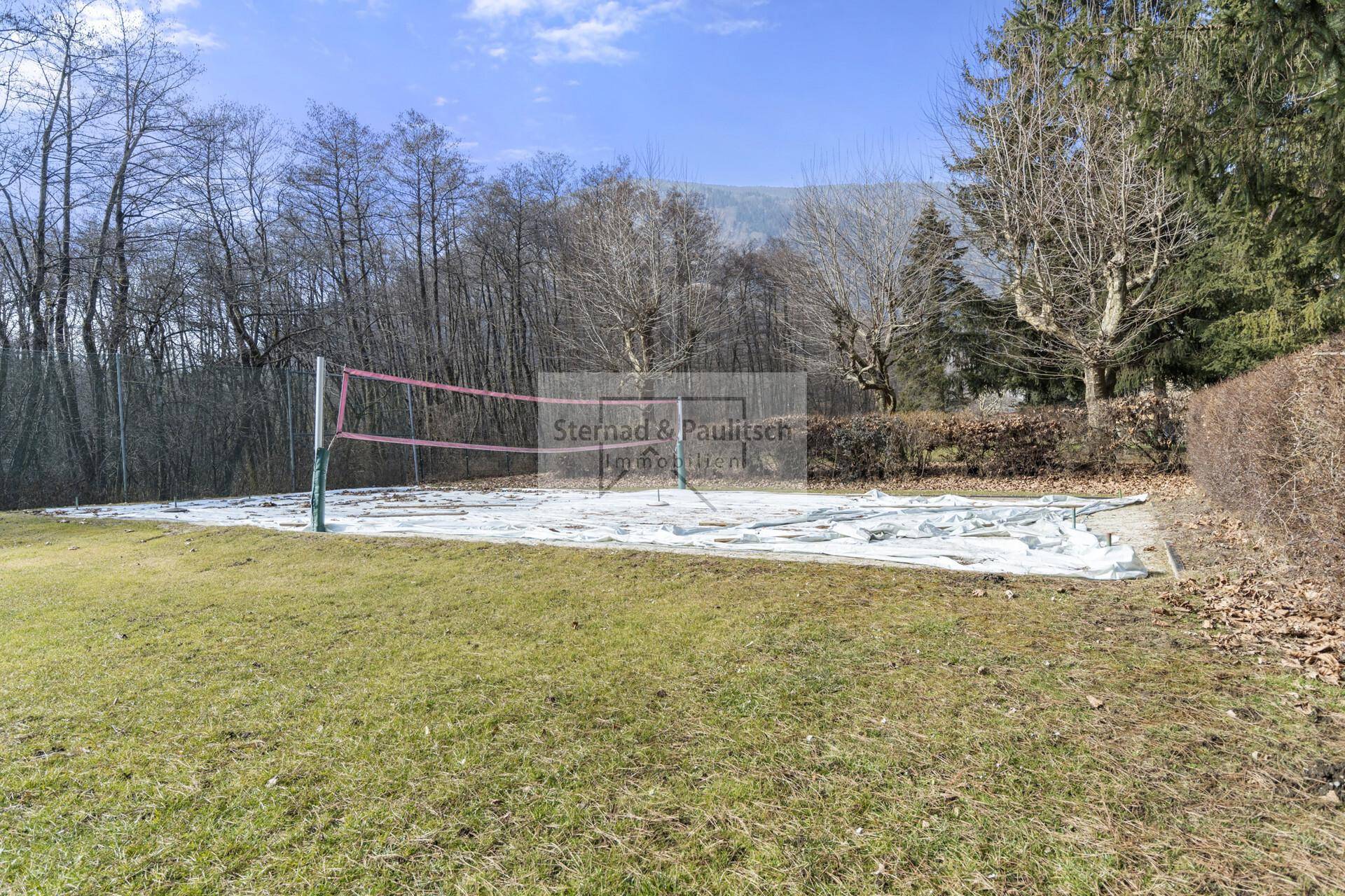 Volleyballfeld auf einer Rasenfläche, umgeben von Bäumen und mit Schnee bedeckt, bereit für den Sommer.