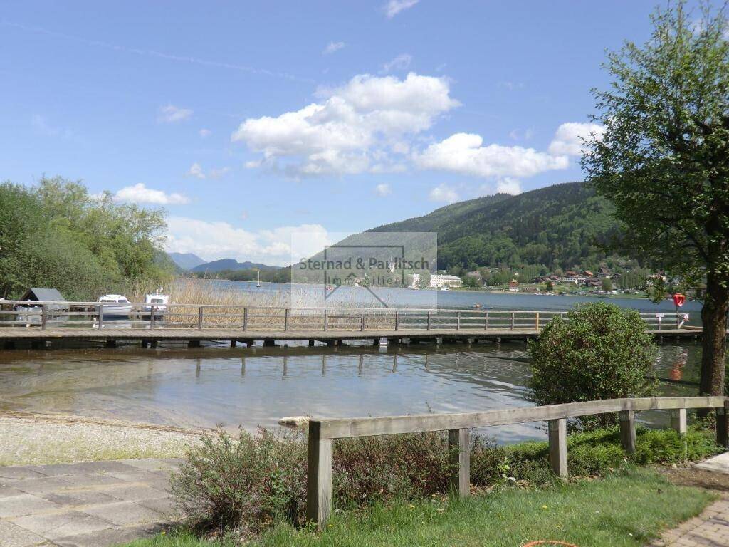 Malerischer Blick auf den See mit langem Holzsteg und bewaldeten Hügeln im Hintergrund an einem sonnigen Tag.
