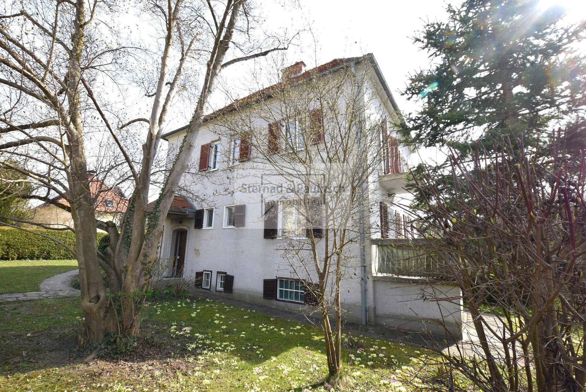 Das Haus von der Seite, umgeben von Bäumen und einem gepflegten Gartenweg.