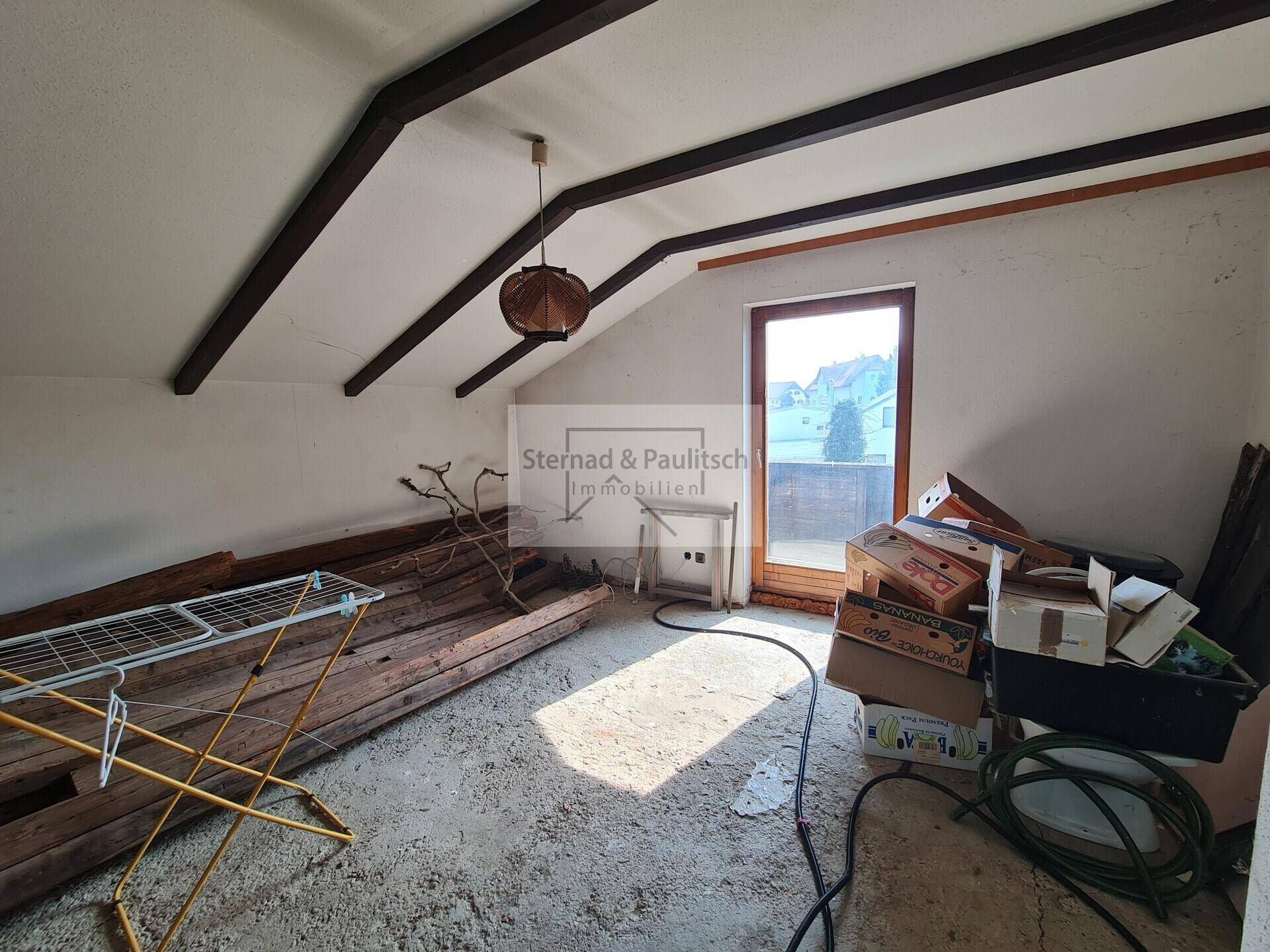 Dachgeschossraum mit Holzbalken und Fenster, genutzt als Abstellfläche mit Renovierungsbedarf.