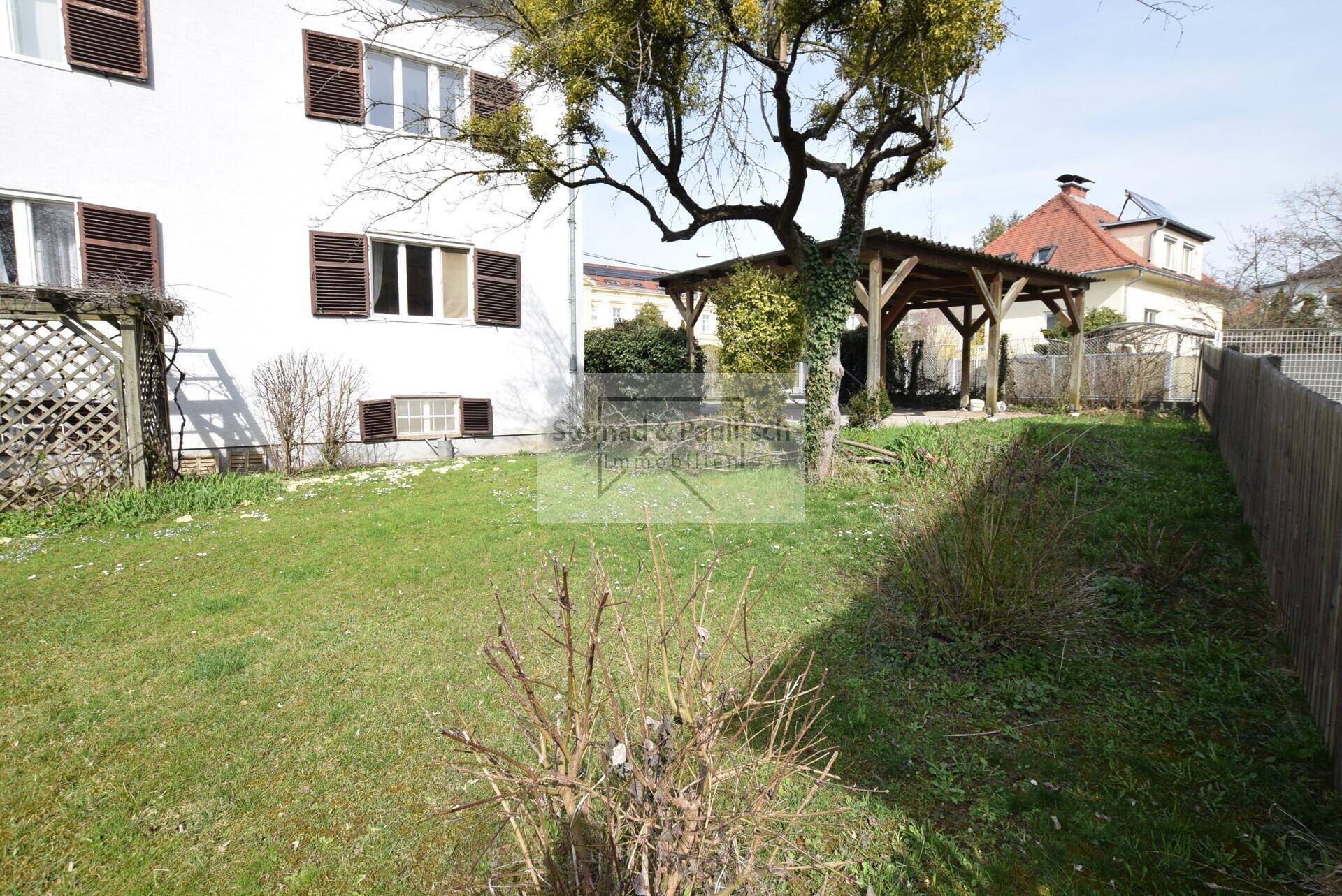 Sonniger Gartenbereich mit Rasenfläche und einer Holzpergola im Hintergrund.
