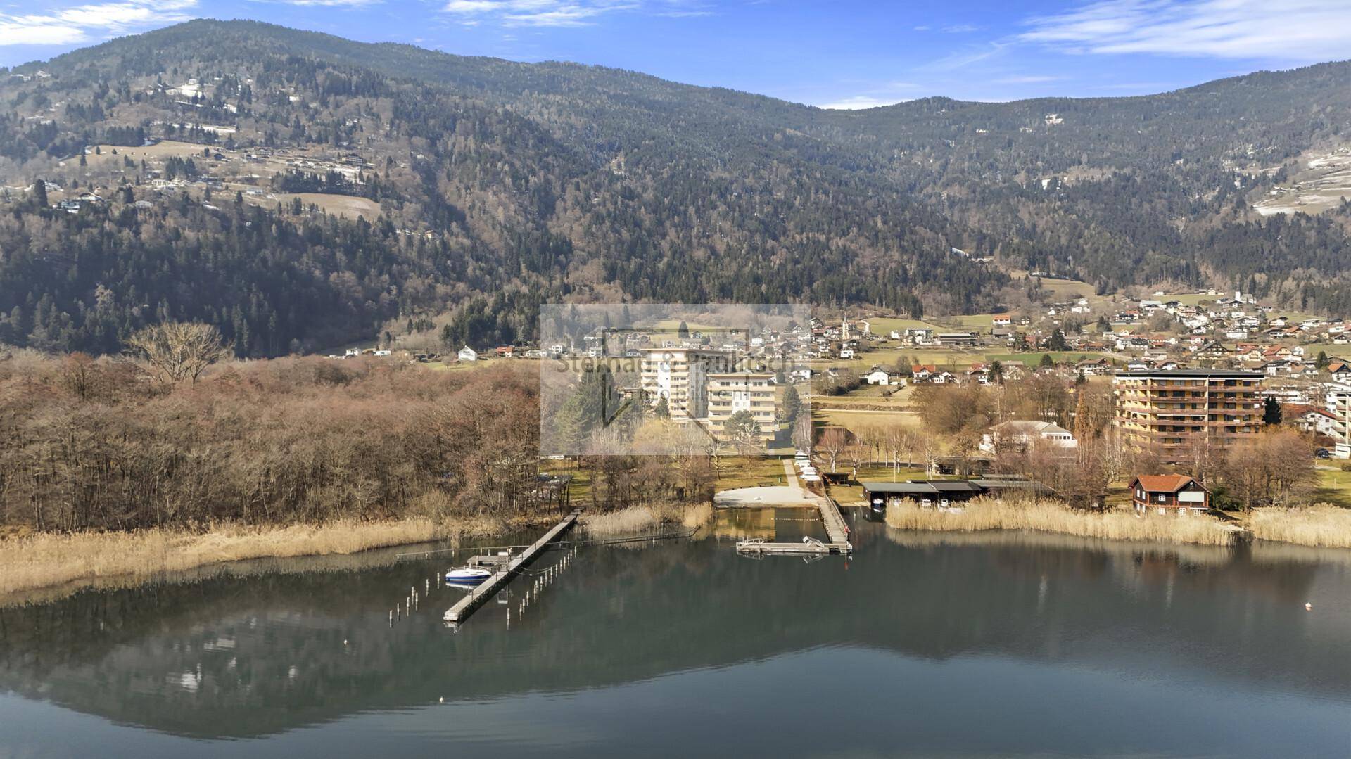Weitläufige Luftaufnahme der Wohnanlage am See mit umliegenden Wäldern und Bergen im Frühling.