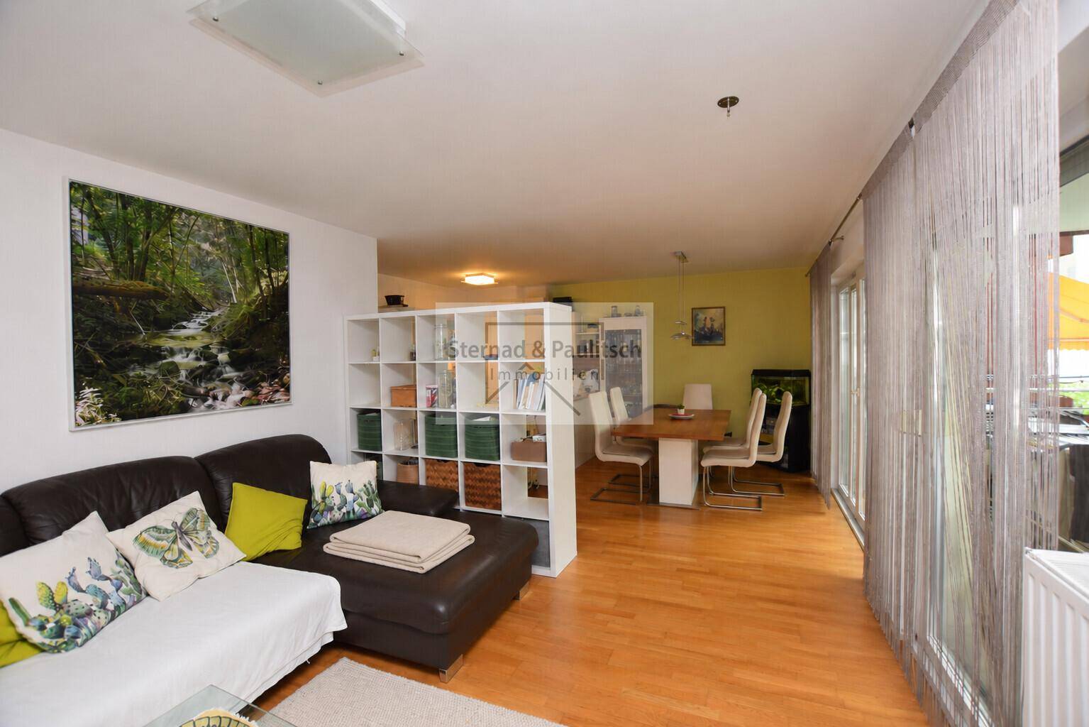 Offener Wohnbereich mit bequemer Couch und Holzboden, verbunden mit dem Essbereich und Balkonzugang.