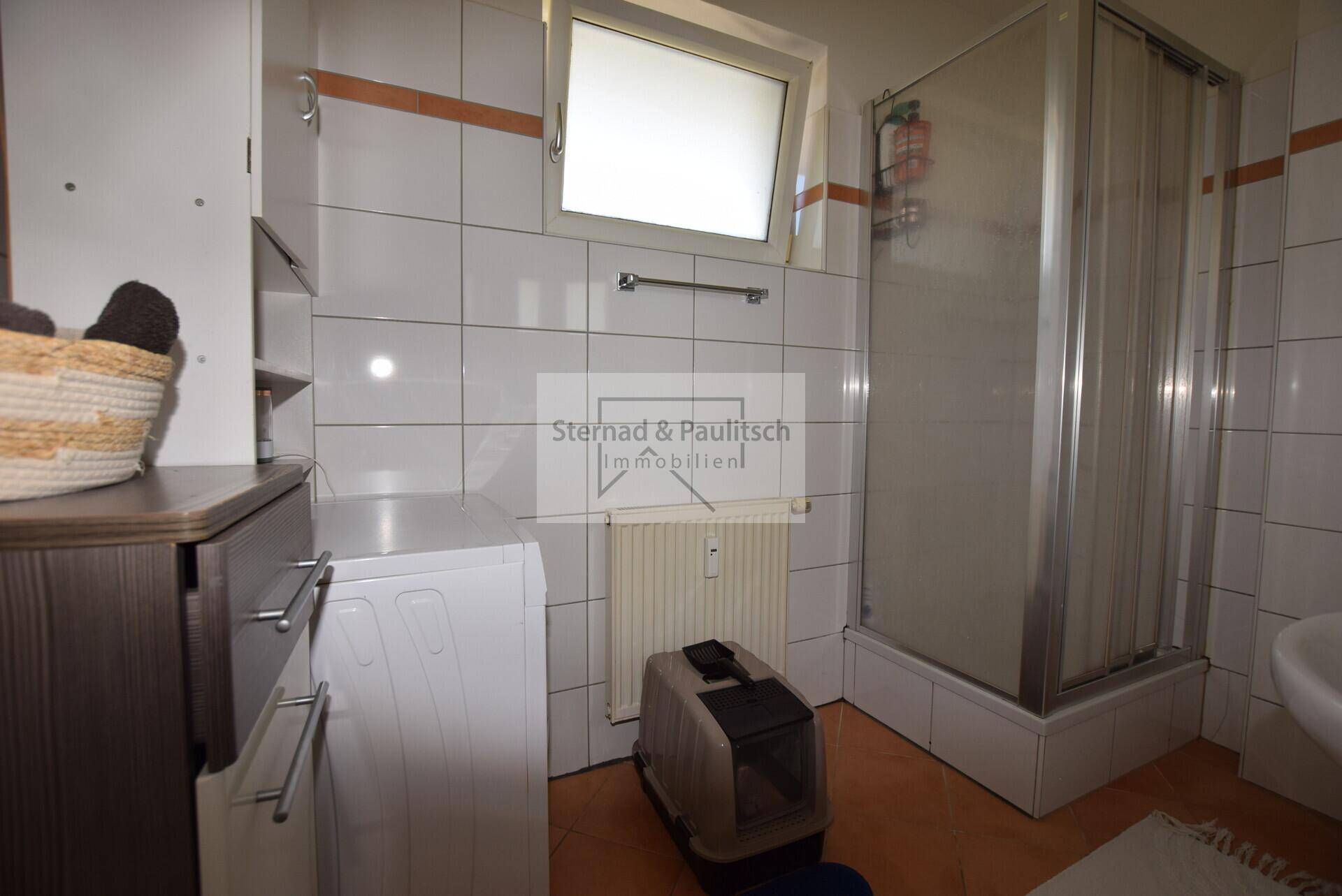 Helles Badezimmer mit Fenster, Duschkabine und praktischem Platz für eine Waschmaschine.
