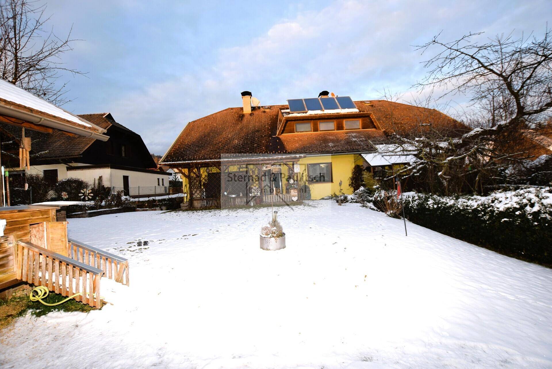 Seitenansicht des Hauses im Winter mit schneebedecktem Garten und Holzanbau.