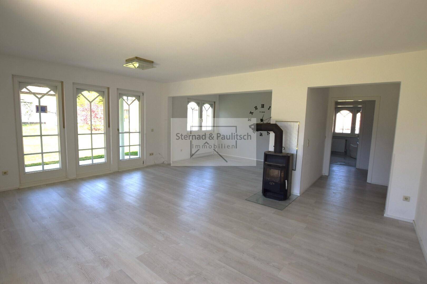 Großzügiges Wohnzimmer mit Kaminofen, hellem Boden und vielen Fenstern für Tageslicht.