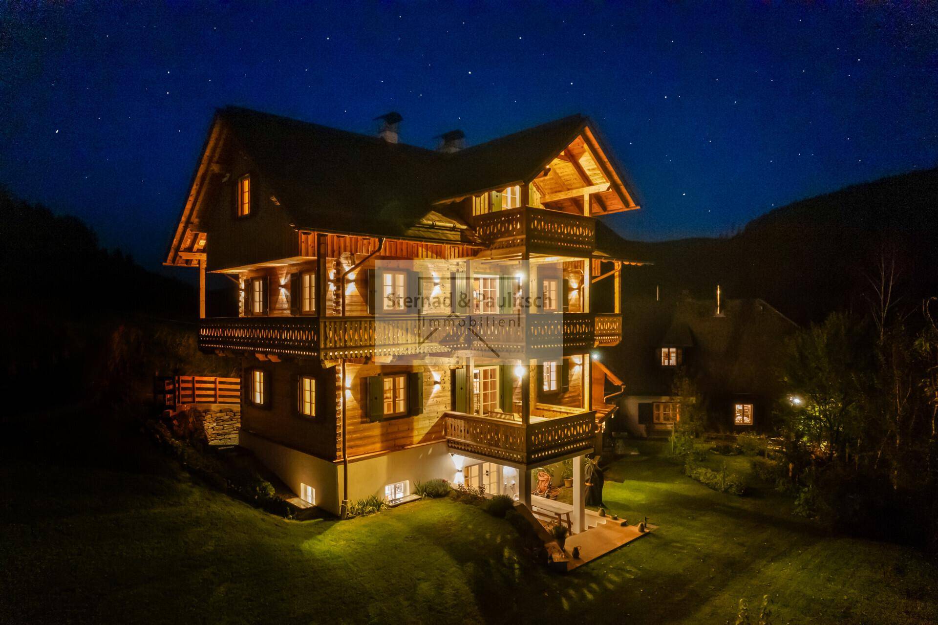 Das beleuchtete Holzchalet bei Nacht unter einem sternenklaren Himmel, eingebettet in die Landschaft.